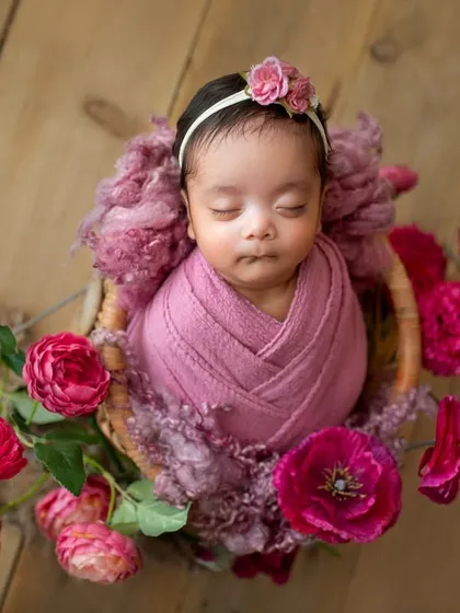 A sweet baby girl nestled in a basket, surrounded by vibrant pink roses. The wood background adds a rustic, warm touch to this floral newborn portrait.