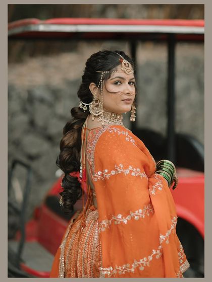 A beautiful back profile of the bride in her orange lehenga, showcasing her intricate hairstyle and jewelry. A modern yet traditional bridal portrait.