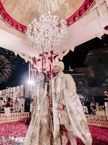 A couple stands together under a grand chandelier within their poolside mandap. This moment, with fireworks in the background, captures the scale and romance of a perfectly executed destination wedding.