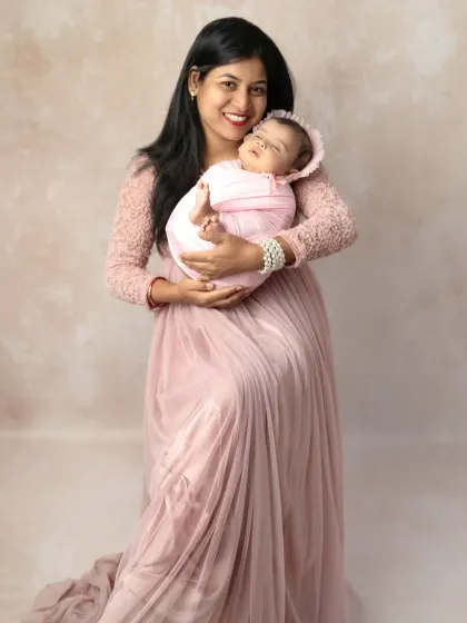 A new mother in a beautiful pink gown sits gracefully, holding her swaddled newborn baby close to her heart in a simple studio setting.