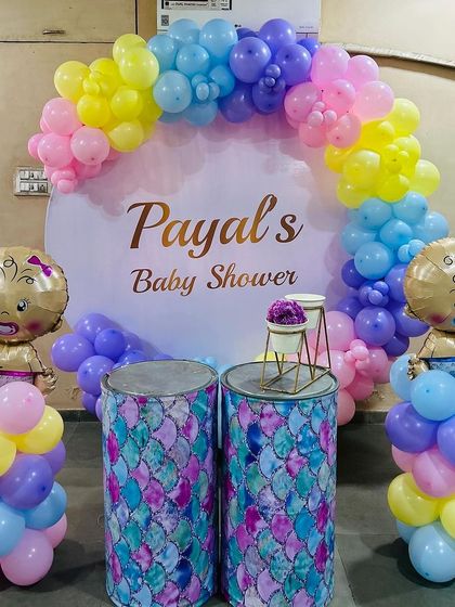 A cheerful baby shower decoration with a mermaid-patterned pedestal and a pastel rainbow balloon arch. The setup is completed with cute baby-shaped foil balloons on columns.