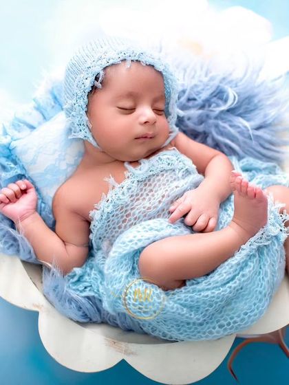 A newborn baby wrapped in a blue swaddle, sleeping peacefully inside a flower-shaped prop against a matching blue floral backdrop.