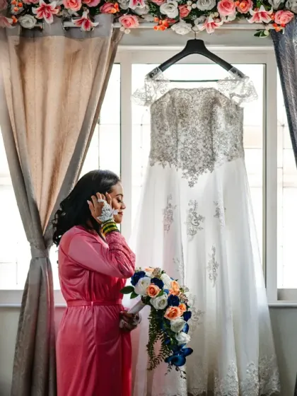 The bride, on the phone, stands next to her wedding dress. A candid moment capturing the modern reality of a wedding day.