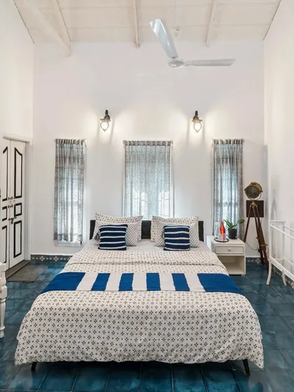 A bright and airy bedroom in a Chikmagalur homestay, with a high vaulted ceiling and blue tiled floors. The simple, clean aesthetic is accented by blue and white block-printed textiles.