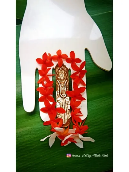 A close-up of the Mataji henna figure, framed by red and white flowers against a green leaf. This composition highlights the delicate details of the Goddess's form, created for the auspicious occasion of Navratri.