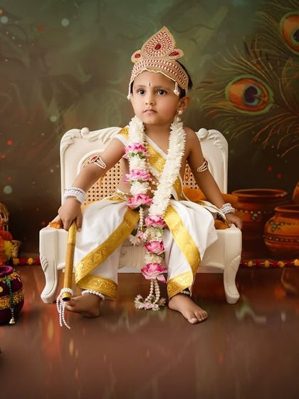 A young boy dressed as Krishna sits majestically on a small white chair, surrounded by peacock feathers and other festive decorations.