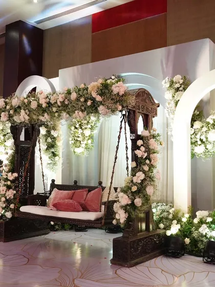 A unique seating arrangement for a ceremony, featuring a traditional wooden swing (jhoola) adorned with white flowers, set against a modern, minimalist backdrop.