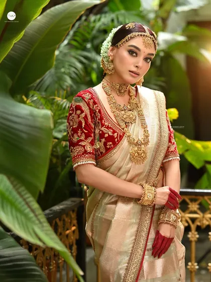 A full-length bridal portrait featuring a stunning off-white and red saree. The lush green background makes the traditional attire pop, and the use of natural light adds a soft, radiant glow to the scene.