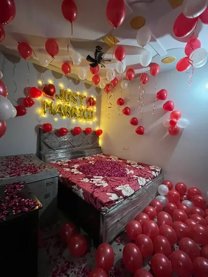 A classic red and white themed 'Just Married' room decoration. This setup includes 'Just Married' foil balloons with warm backlighting, a bed covered in a rose petal heart, and the floor filled with balloons.