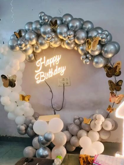 A chic and simple first birthday setup. A circular balloon arch in silver and white is accented with butterfly details and a 'Happy Birthday' neon sign, complemented by a light-up number '1'.