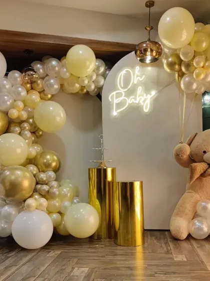 This 'Oh Baby' shower setup is a popular choice, featuring a soft and elegant color palette of white, cream, and chrome gold balloons. The arrangement is built around a white arched backdrop with a neon sign and includes a large teddy bear prop and golden pedestals for a beautiful display.