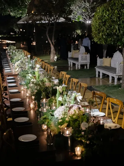 A long-table welcome dinner setup at night, illuminated by candlelight. The focus on intimate lighting and lush green centerpieces creates a warm and inviting atmosphere for guests to connect.
