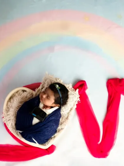 The word 'LOVE' is spelled out with red fabric, with the baby nestled inside the 'O' as a heart. A painted rainbow in the background adds a touch of whimsy.