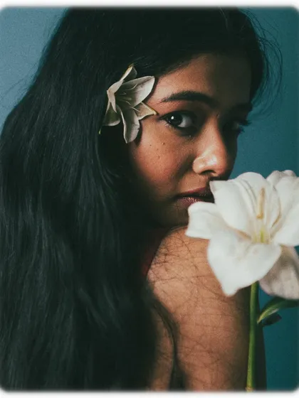 This composition uses two flowers to frame the model's face. Her expression is soft and direct, creating a beautiful contrast between the delicate props and her engaging gaze. The lighting is soft, enhancing the dreamy quality of the image.