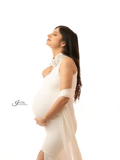 A serene solo portrait. The expecting mother, dressed in a simple white gown, looks up with her eyes closed, creating a moment of quiet reflection during her pregnancy journey.