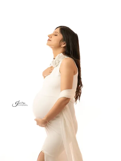 A serene solo portrait. The expecting mother, dressed in a simple white gown, looks up with her eyes closed, creating a moment of quiet reflection during her pregnancy journey.