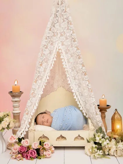 A baby sleeps peacefully in a miniature bed under a delicate lace tent, surrounded by candles and flowers. This setup is perfect for a romantic and whimsical newborn photoshoot.