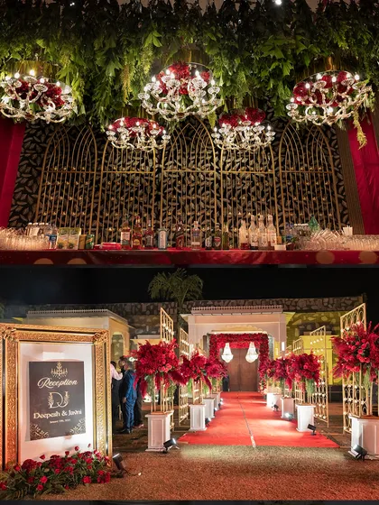 The reception bar was a masterpiece of design, set against a golden jaali backdrop with hanging chandeliers adorned with red roses. The entrance featured a red carpet and grand floral arrangements.