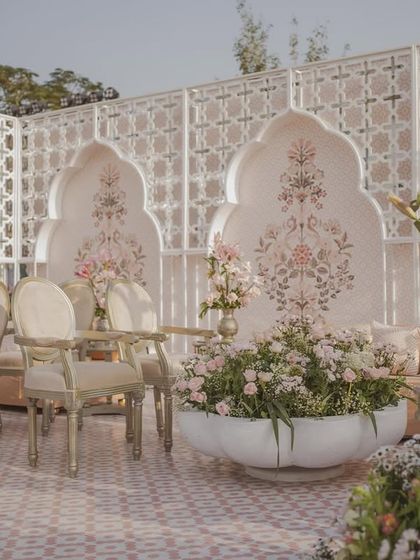 The main stage for a pastel-themed Mughal wedding. Ornate chairs are set against a backdrop of hand-painted arches, flanked by large bowls of fresh flowers.