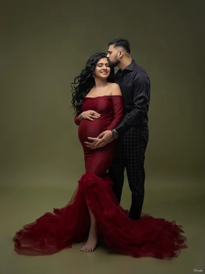 A tender moment in the studio. He gently kisses her forehead as she cradles her bump, both dressed in elegant, coordinating outfits.