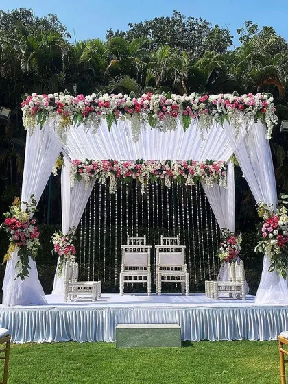 This is an elegant vedi mandap I designed for an outdoor day wedding. The structure is draped in clean white fabric, with lush floral arrangements of pink and white roses on top and along the pillars, creating a modern yet timeless look for the pheras.