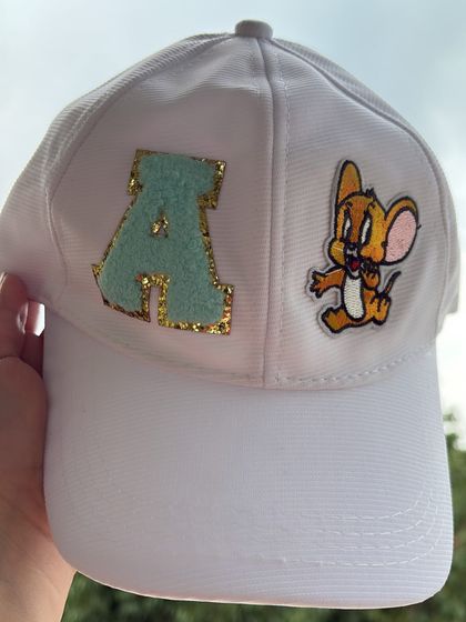 A detailed look at a finished white cap personalized with a glittery chenille 'A' patch and a classic Jerry Mouse character patch. This showcases the clean application and fun combinations possible.