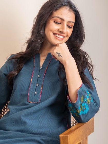 A smiling model in our Haveli Crewel Blue Kurta. The stylized neckline and intricate embroidery on the cuffs make it a standout piece for any occasion.