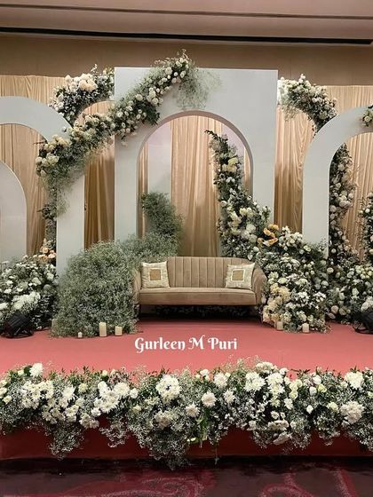 A full stage setup using the modern white arch concept. The arches are layered to create a sense of depth, with a simple couch at the center. The entire stage is framed by a profusion of white flowers and baby's breath for a soft, romantic look.