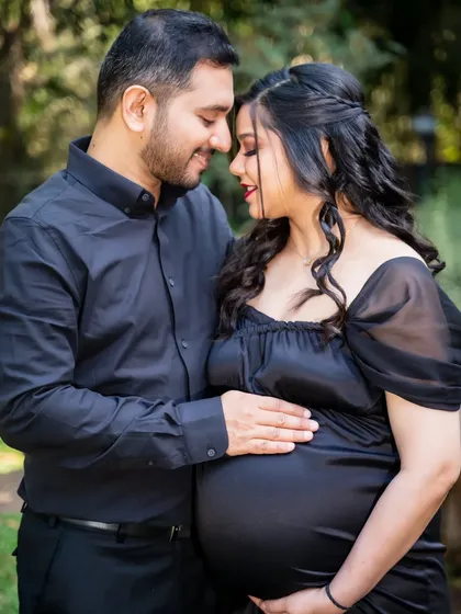 An intimate close-up of a couple, both dressed in black, sharing a quiet moment. The focus is on their connection and the baby bump they cradle together.