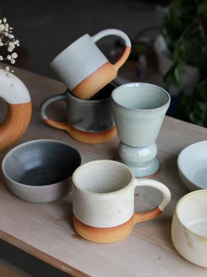 An elegant collection of mugs, bowls, and a donut vase in muted tones, showcasing clean forms and the beauty of the natural clay body.