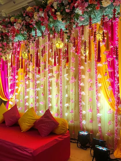 A classic Haldi or Mehendi stage with a backdrop of bright yellow and pink drapes and strings of hanging flowers, creating a festive and joyful setting.