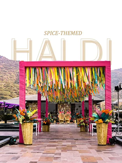 The grand entrance to the spice-themed Haldi, with a vibrant pink frame and a ceiling of colorful ribbons, set against the beautiful backdrop of the Aravalli hills in Udaipur.