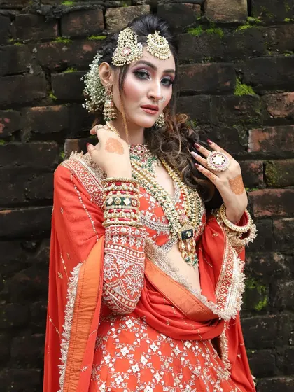 A stunning outdoor shot of a bride in an orange lehenga. Her makeup features dramatic eyes and is paired with a messy, romantic hairstyle.