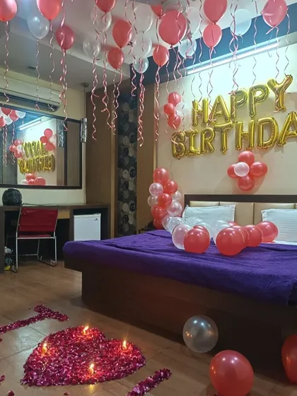 A hotel room decorated for a birthday surprise. The setup includes red and white balloons, "Happy Birthday" foils, and a rose petal heart on the floor with candles.