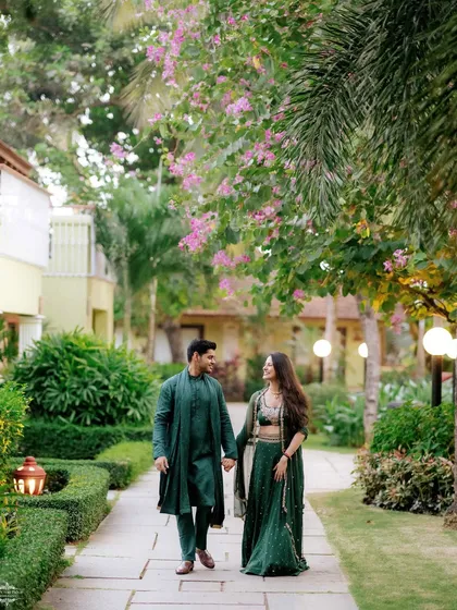 A couple in matching deep green traditional wear walks hand-in-hand through the lush gardens of the Novotel in Goa.