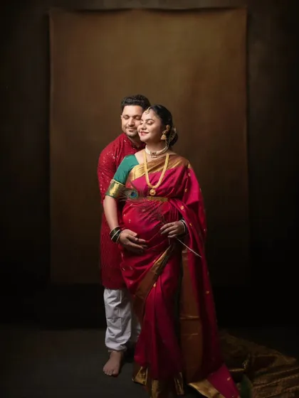 A full-length portrait showcasing the beauty of a traditional maternity look. The deep red saree drapes beautifully over her baby bump, creating an image of strength, grace, and cultural pride.