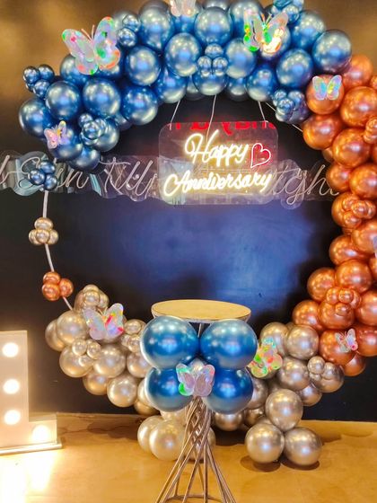 Celebrate your first year together with a special decoration. This first-anniversary setup features a balloon ring with a mix of blue, copper, and silver chrome balloons, butterfly cutouts, a "Happy Anniversary" neon sign, and a light-up number "1".