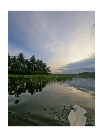 A first-person perspective from a kayak, showing the paddle dipping into the water and the beautiful sky above.