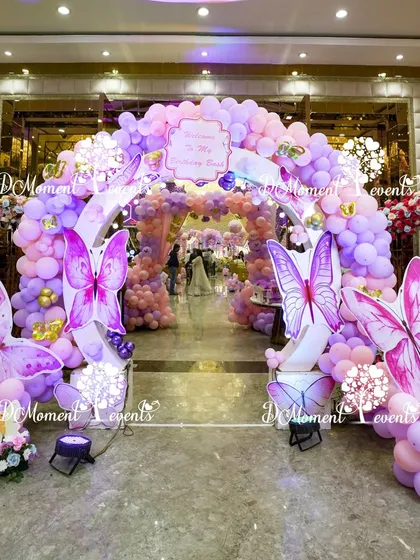 A grand butterfly-themed entrance archway for Aarna's first birthday. This elaborate setup features a tunnel of purple and pink balloons, accented with large, detailed butterfly wing props for a spectacular welcome.