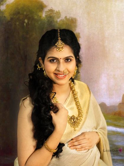 A close-up portrait inspired by the art of Raja Ravi Varma. The mother-to-be, adorned in gold jewelry and a cream saree, smiles warmly against a soft, painted background.