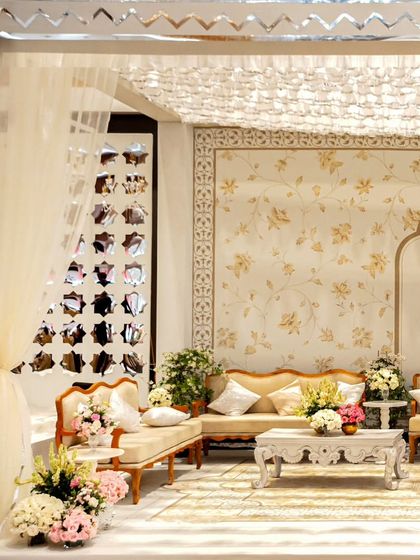 A side view of the elegant white and gold lounge. The space is defined by a mirrored frame with a zigzag pattern, soft drapes, and a mix of classic furniture and fresh floral arrangements.