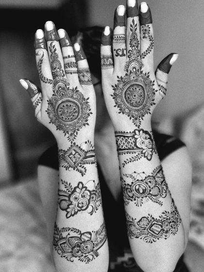 A black and white photo highlighting the intricate patterns of an engagement mehendi. The design includes mandala art on the palms and floral bands on the arms.
