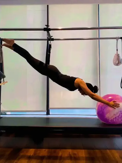 A client demonstrates a full-body stretch on the Cadillac, reaching for the stability ball. This move requires both strength to hold the position and flexibility to achieve the extension.