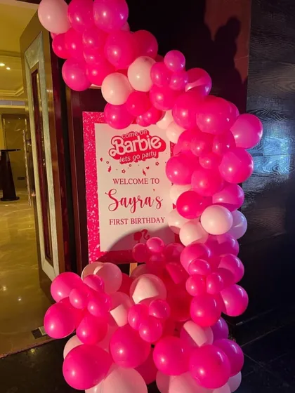 A Barbie-themed entrance decoration with a pink balloon arch and a personalized welcome sign for a first birthday party.
