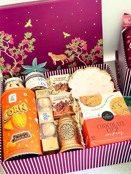 A Box of Delicacy, this hamper comes in a stunning purple festive box. It's overflowing with treats like Blue Tokai cookies, a lotus urli candle, a festive lantern, crunchy cheese corn, and premium nuts, offering something for everyone.