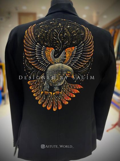 The back of a black blazer featuring a detailed, hand-painted falcon with spread wings. The use of gold and bronze tones gives the artwork a metallic, shimmering effect.