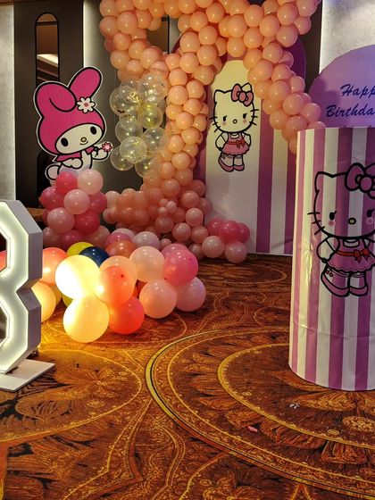 A close-up of the Hello Kitty side of the party stage, with a large balloon number '8', character cutouts, and a pink-and-white striped theme plinth.