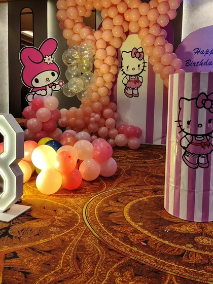 A close-up of the Hello Kitty side of the party stage, with a large balloon number '8', character cutouts, and a pink-and-white striped theme plinth.
