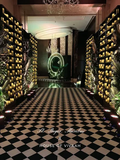 The entrance to a bold cocktail night at St. Regis Mumbai. A black-and-white checkerboard floor leads guests down an aisle of candle walls and tropical foliage, ending at a glowing neon art piece.