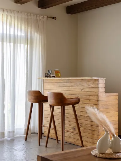A simple wooden bar counter with modern high chairs provides a casual spot for drinks. The sheer curtains diffuse the natural light, adding to the room's soft and breezy atmosphere.